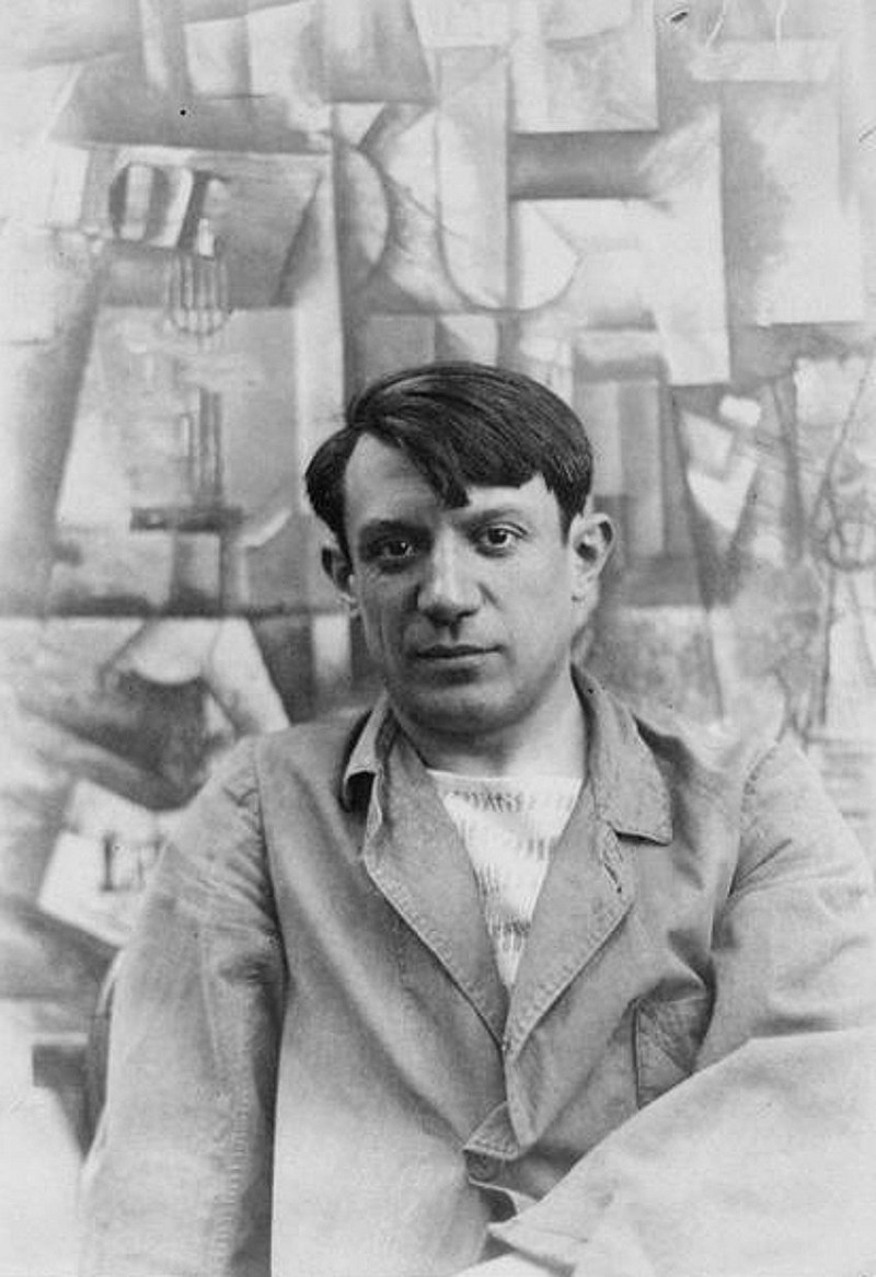 Portrait photograph of Pablo Picasso in front of his painting The Aficionado (Kunstmuseum Basel) at Villa les Clochettes, Sorgues, France, summer 1912; Anonymous Unknown author, Public domain, via Wikimedia Commons